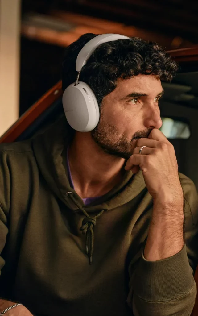 A person wearing Sonos Ace headphones while relaxing, with a JBL Flip 7 portable speaker visible in the background, showcasing versatile audio gifts.
