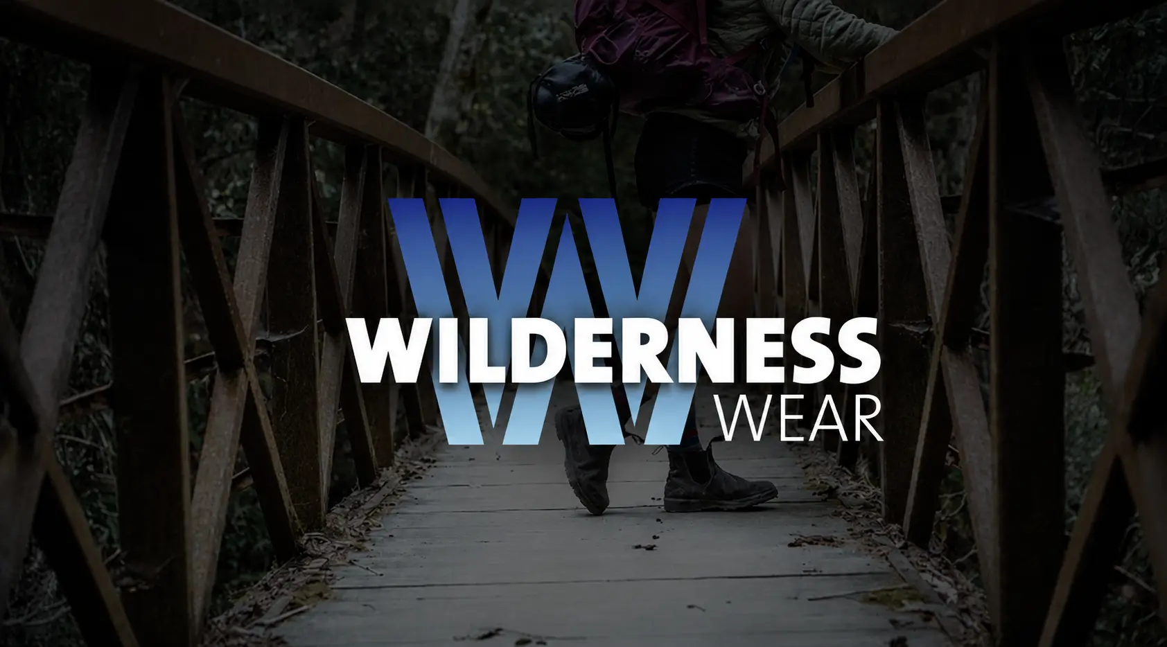 A hiker walks across a wooden bridge in the forest, featuring the Wilderness Wear brand logo overlaid in the center of the image.